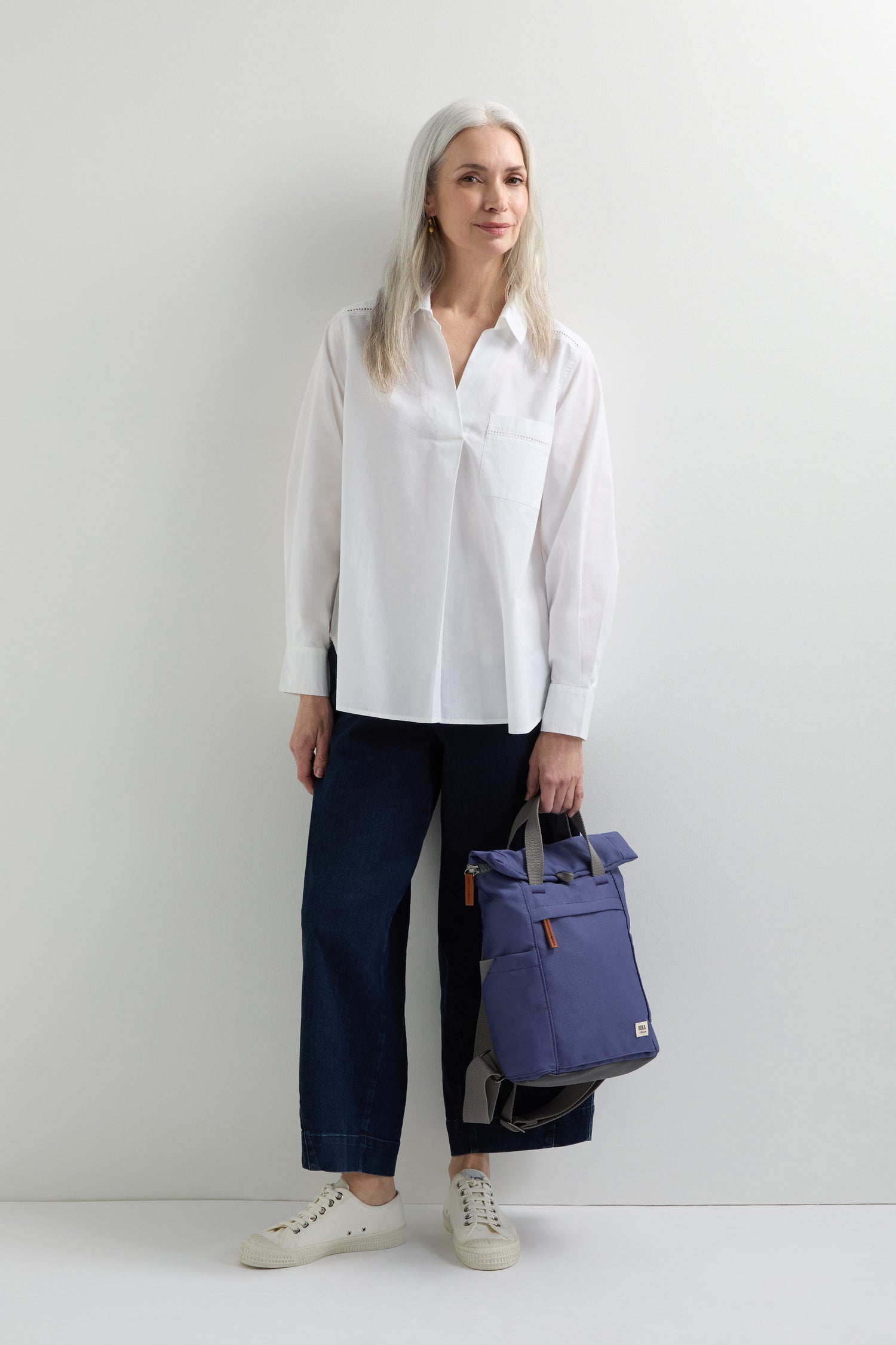 A woman with long gray hair stands against a white wall wearing a white blouse, dark pants, and white sneakers. She holds the Recycled Canvas Small Finchley Bag, showcasing her commitment to sustainable fashion with this eco-conscious choice.