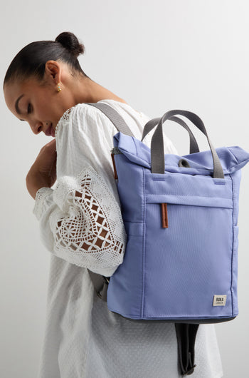 A person with a light blouse and an eco-conscious blue Recycled Canvas Small Finchley Bag smiles while looking down, standing against a plain background.