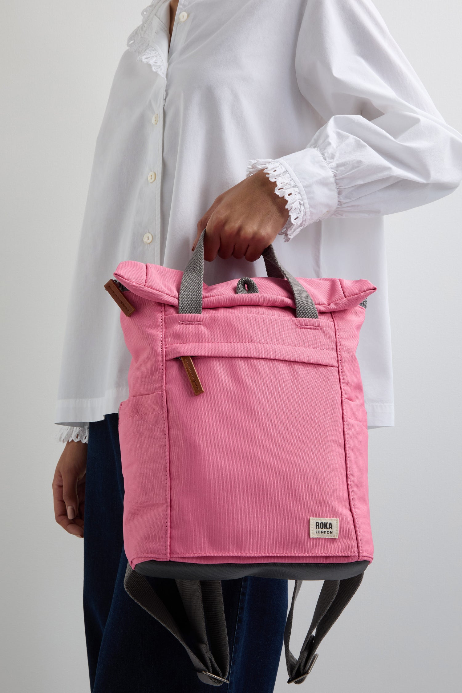 A person holds a pink Recycled Canvas Small Finchley Bag, wearing a white blouse and dark pants, highlighting its eco-conscious design ideal for sustainable fashion enthusiasts.