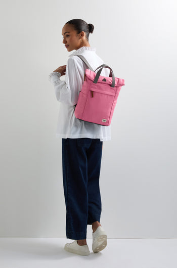 A person in a white blouse and dark pants, with hair tied back, stands against a plain background carrying a pink Recycled Canvas Small Finchley Bag.