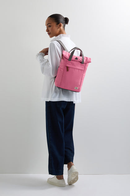 A person in a white blouse and dark pants, with hair tied back, stands against a plain background carrying a pink Recycled Canvas Small Finchley Bag.