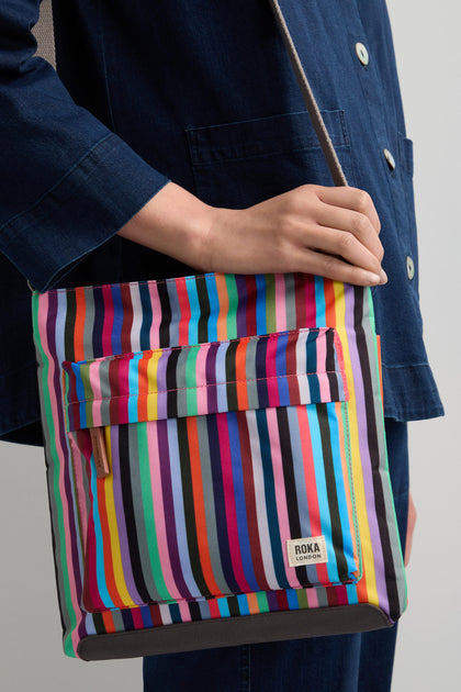 A person carries the Multi Stripe Recycled Canvas Kennington Bag, a stylish shoulder bag with vertical multicolored stripes and a front pocket. The "ROKA London" label complements their dark blue outfit perfectly.