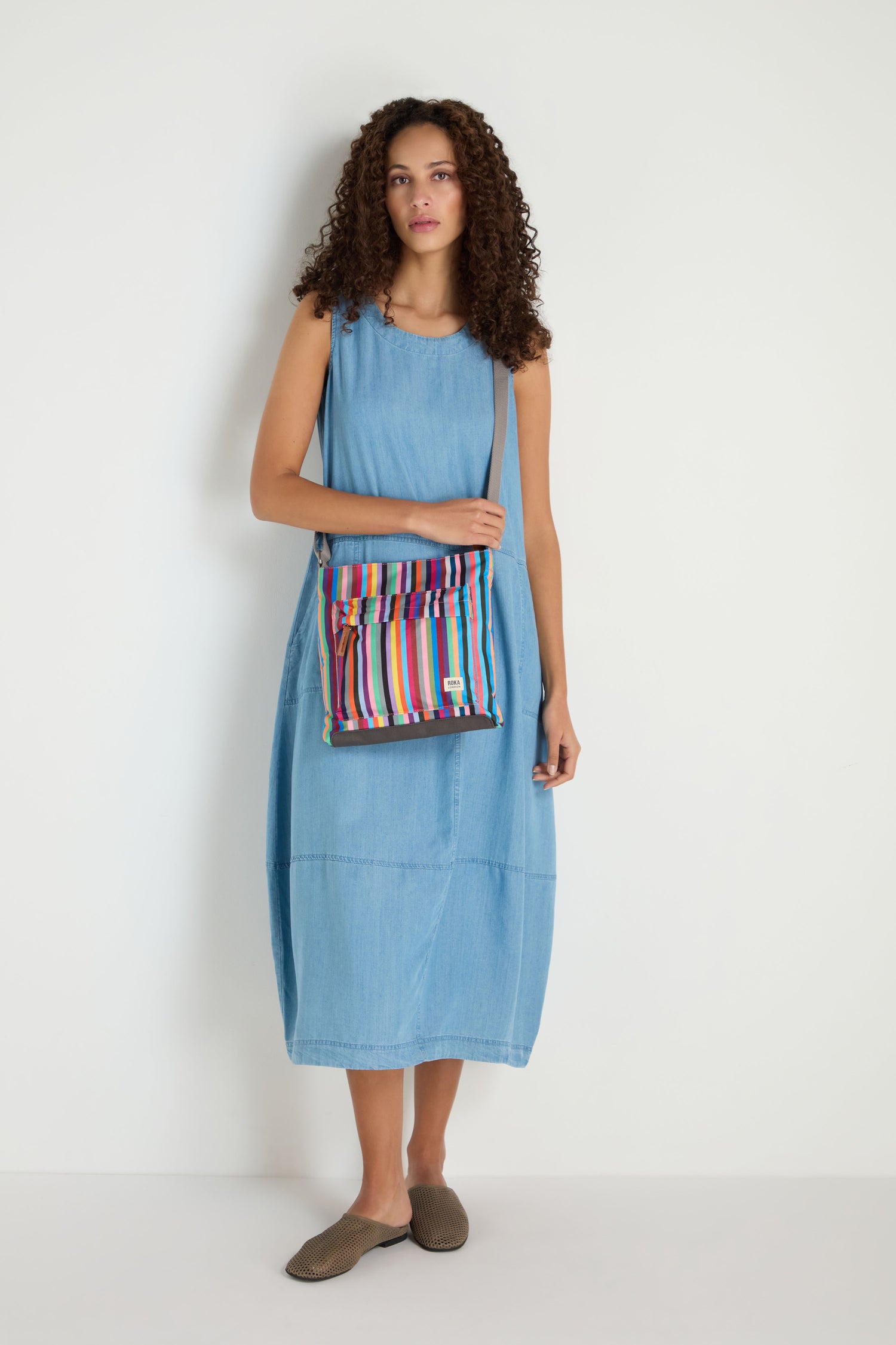 A woman in a sleeveless light blue denim dress stands against a white wall, wearing brown slip-on shoes and carrying the Recycled Canvas Kennington Bag.