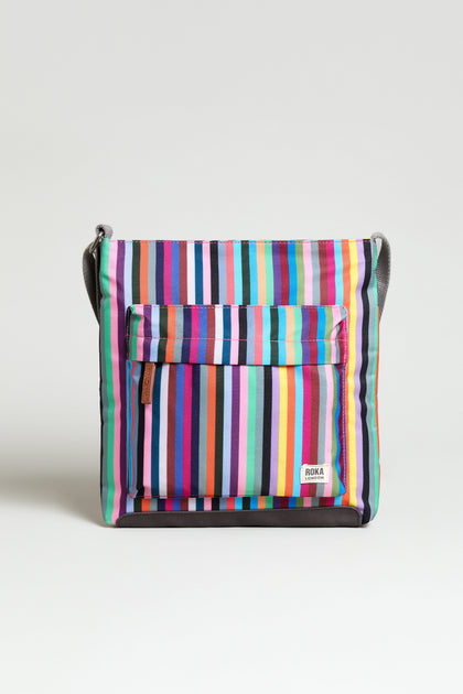 The Recycled Canvas Kennington Bag is a sustainable crossbody with a front pocket, multicolored vertical stripes, and a small "ROKA" label on the lower right corner, crafted from recycled canvas.