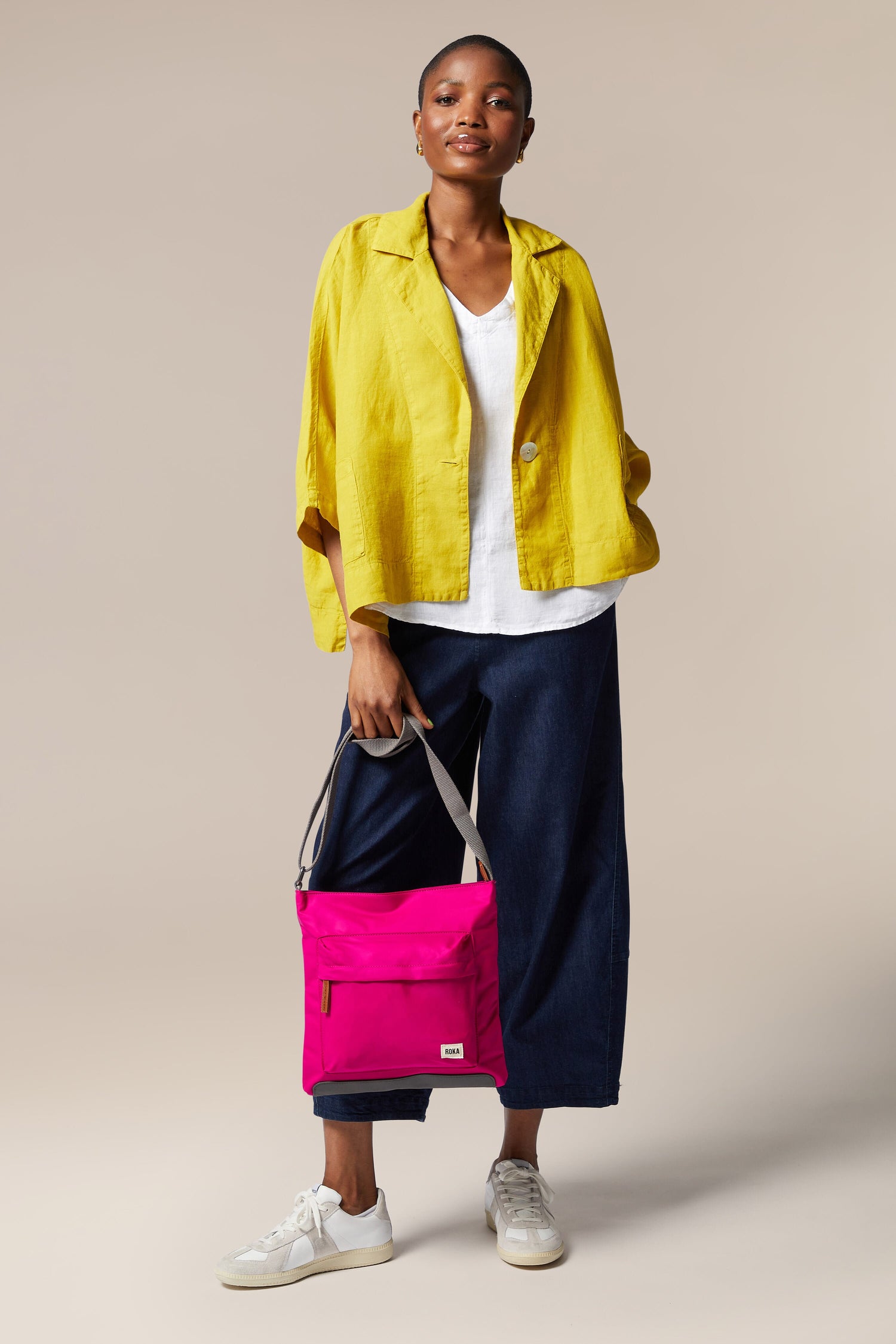 A woman in a pink jacket and yellow pants is holding a sleek Kennington Crossbody Bag.