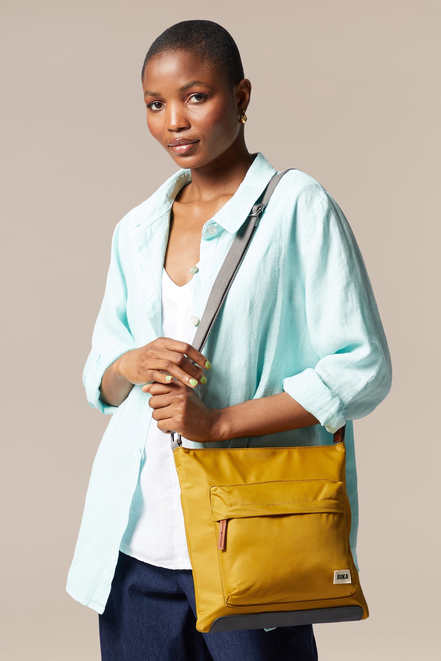 A woman is holding a sleek, vegan yellow Kennington Crossbody Bag with an adjustable strap.