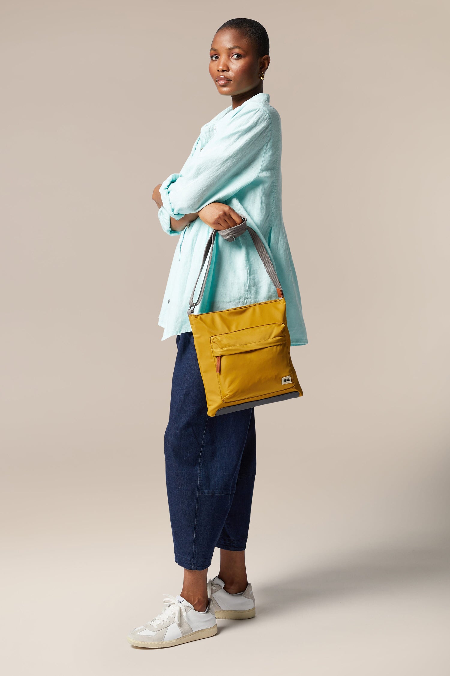 A woman in a blue shirt and jeans is holding a Kennington Crossbody Bag with an adjustable strap.