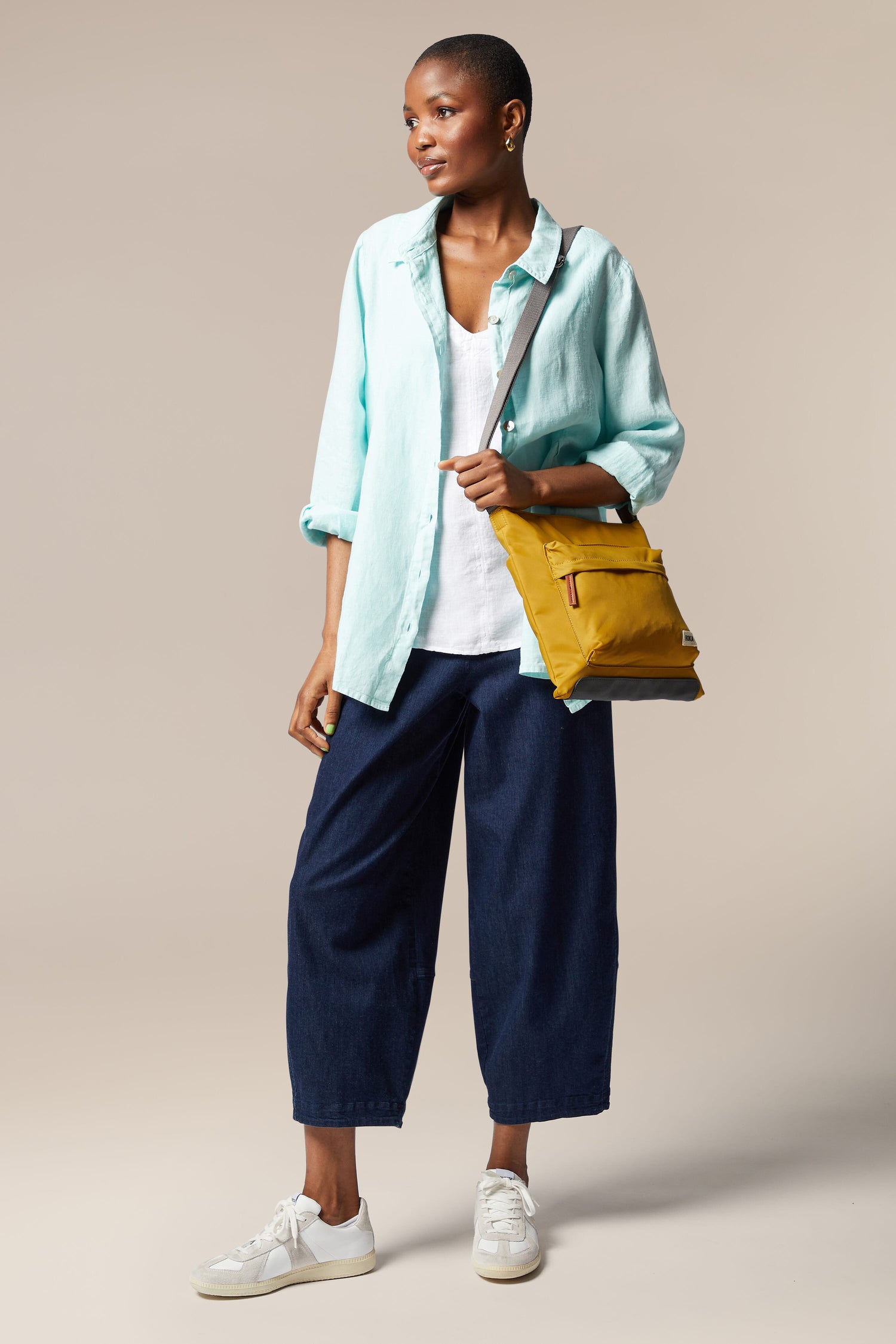 A woman in a sleek blue shirt and blue pants is holding a Kennington Crossbody Bag with an adjustable strap.