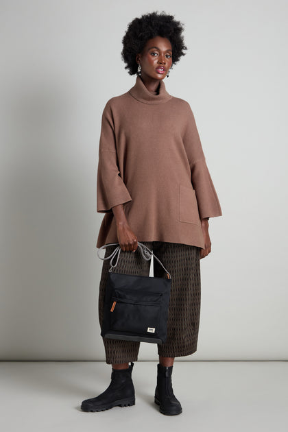 A person stands against a plain backdrop wearing a brown turtleneck, patterned pants, and black boots, holding the stylish Recycled Nylon Kennington Crossbody Bag.