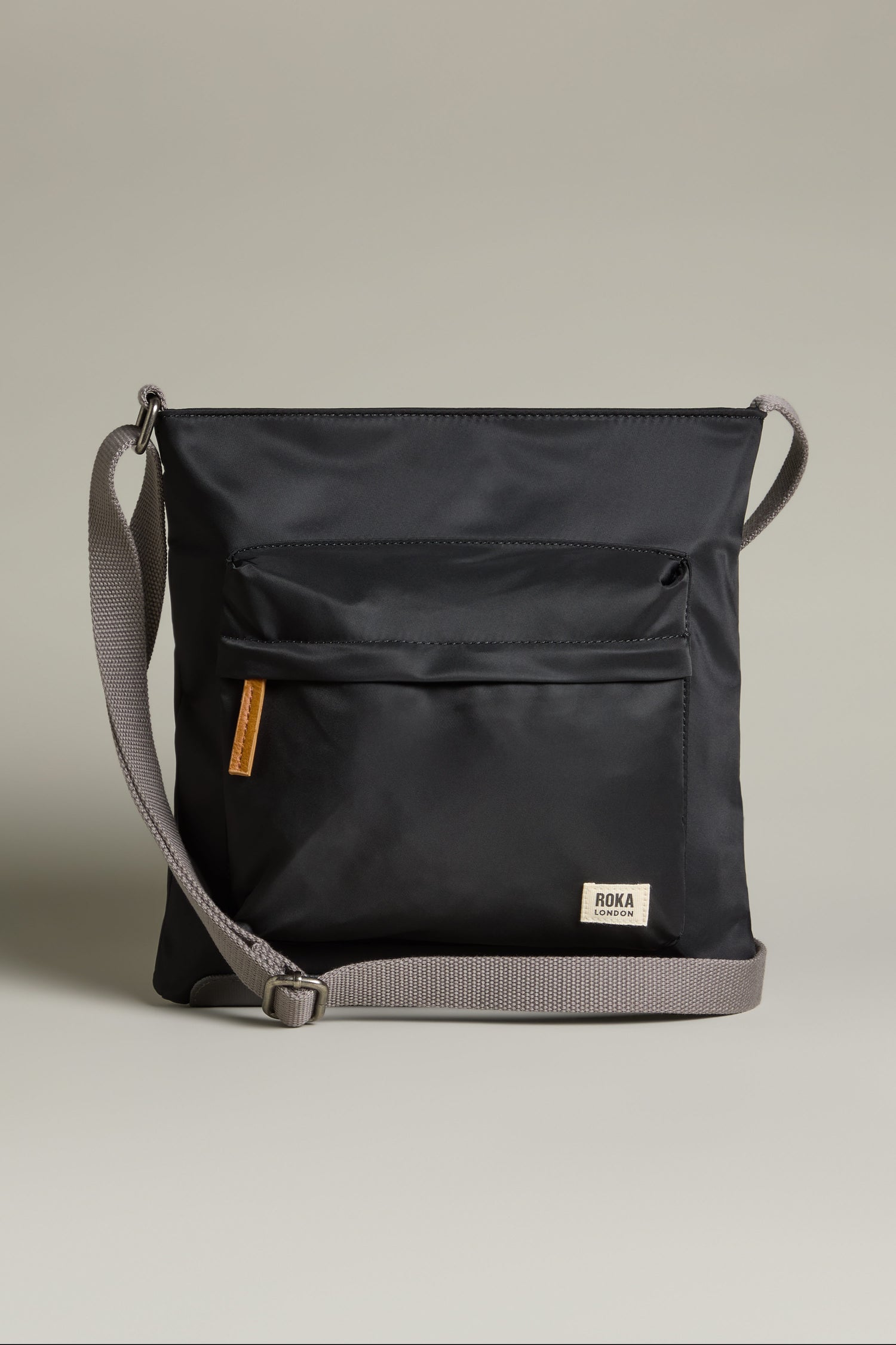 The Recycled Nylon Kennington Crossbody Bag is a chic black bag made from water-resistant fabric, showcasing a front pocket and an adjustable gray strap, set against a simple backdrop.