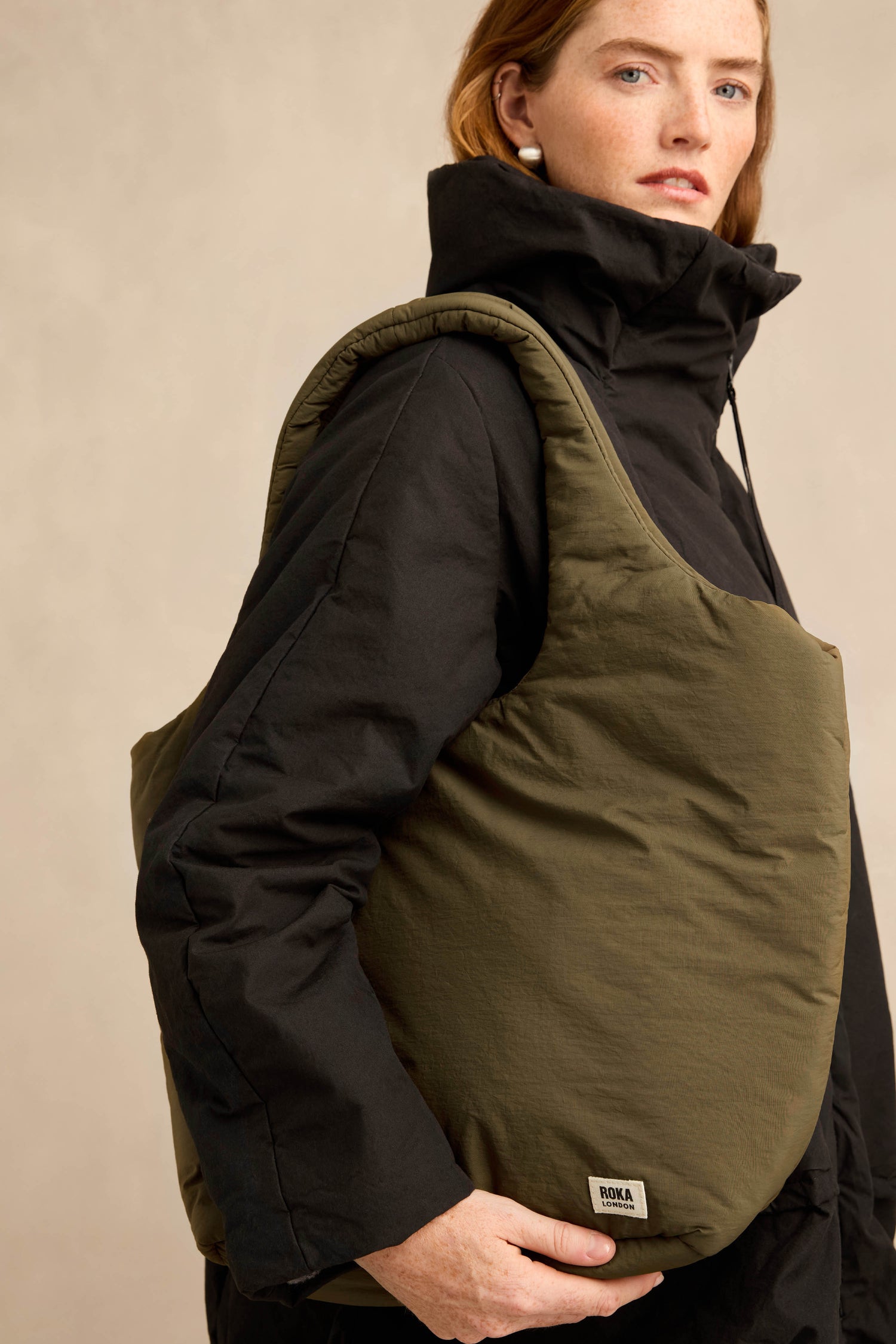A person wearing a black jacket carries an oversized Oxford Moss Shoulder Bag in olive green, featuring padded detailing and a small visible label on the bottom corner.