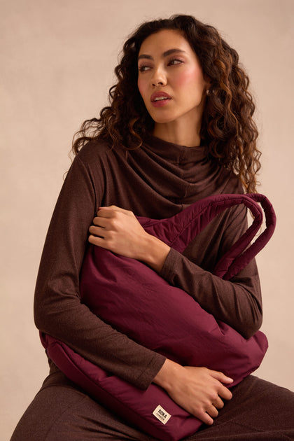 A woman with long curly hair, dressed in a brown outfit, sits holding the Oxford Merlot Shoulder Bag—a large maroon quilted bag featuring a padded laptop sleeve—against a plain beige background.