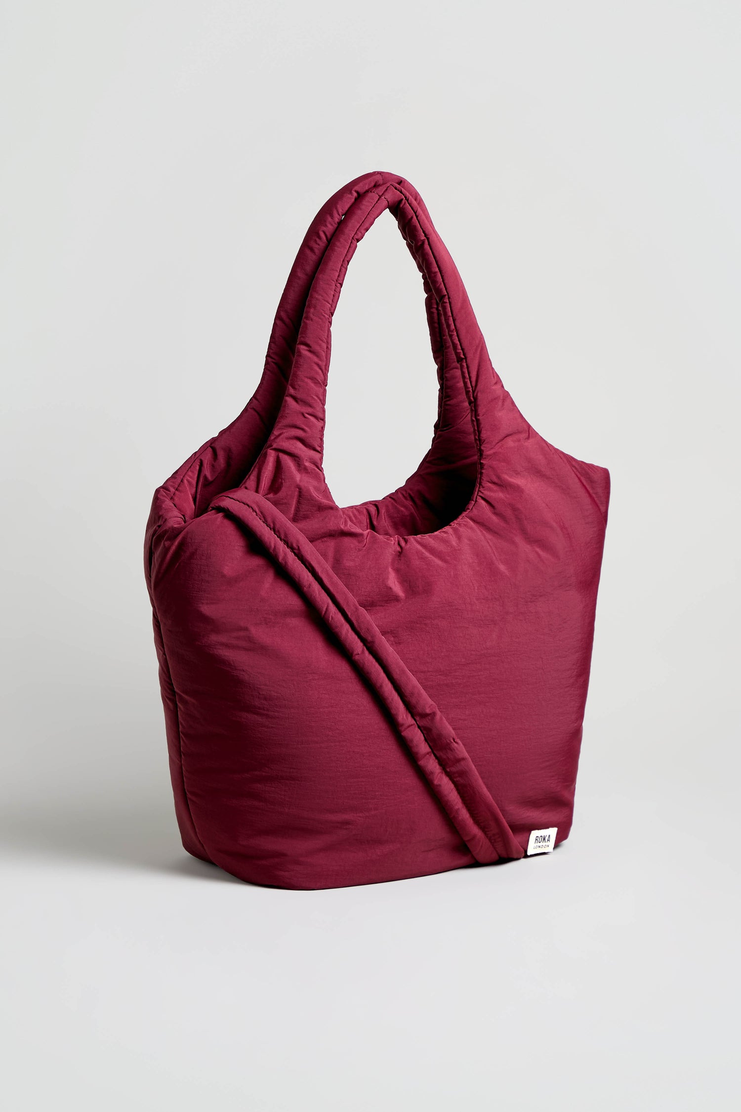 The Oxford Merlot Shoulder Bag is a padded, deep red tote with a soft, quilted texture and two handles, standing upright against a plain light background. This weather-resistant bag blends stylish design with practicality for everyday use.