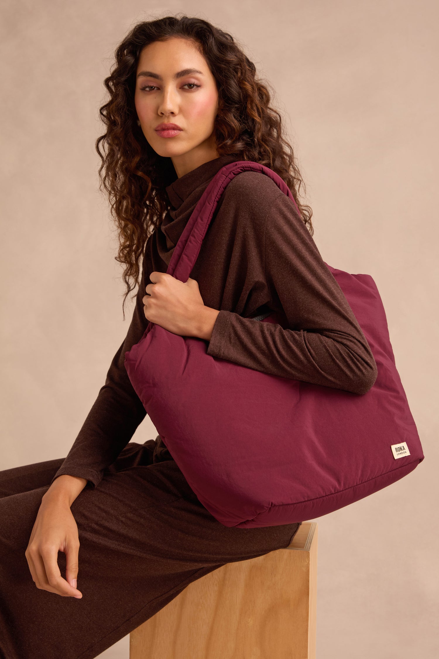 A woman in a brown outfit sits on a wooden stool, holding the Oxford Merlot Shoulder Bag with a padded laptop sleeve on her shoulder against a neutral background.