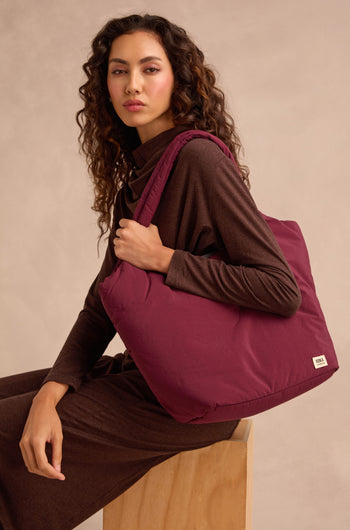 A woman in a brown outfit sits on a wooden stool, holding the Oxford Merlot Shoulder Bag with a padded laptop sleeve on her shoulder against a neutral background.