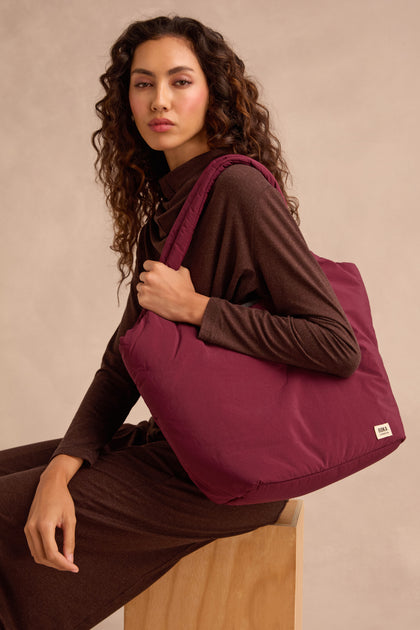 A woman in a brown outfit sits on a wooden stool, holding the Oxford Merlot Shoulder Bag with a padded laptop sleeve on her shoulder against a neutral background.