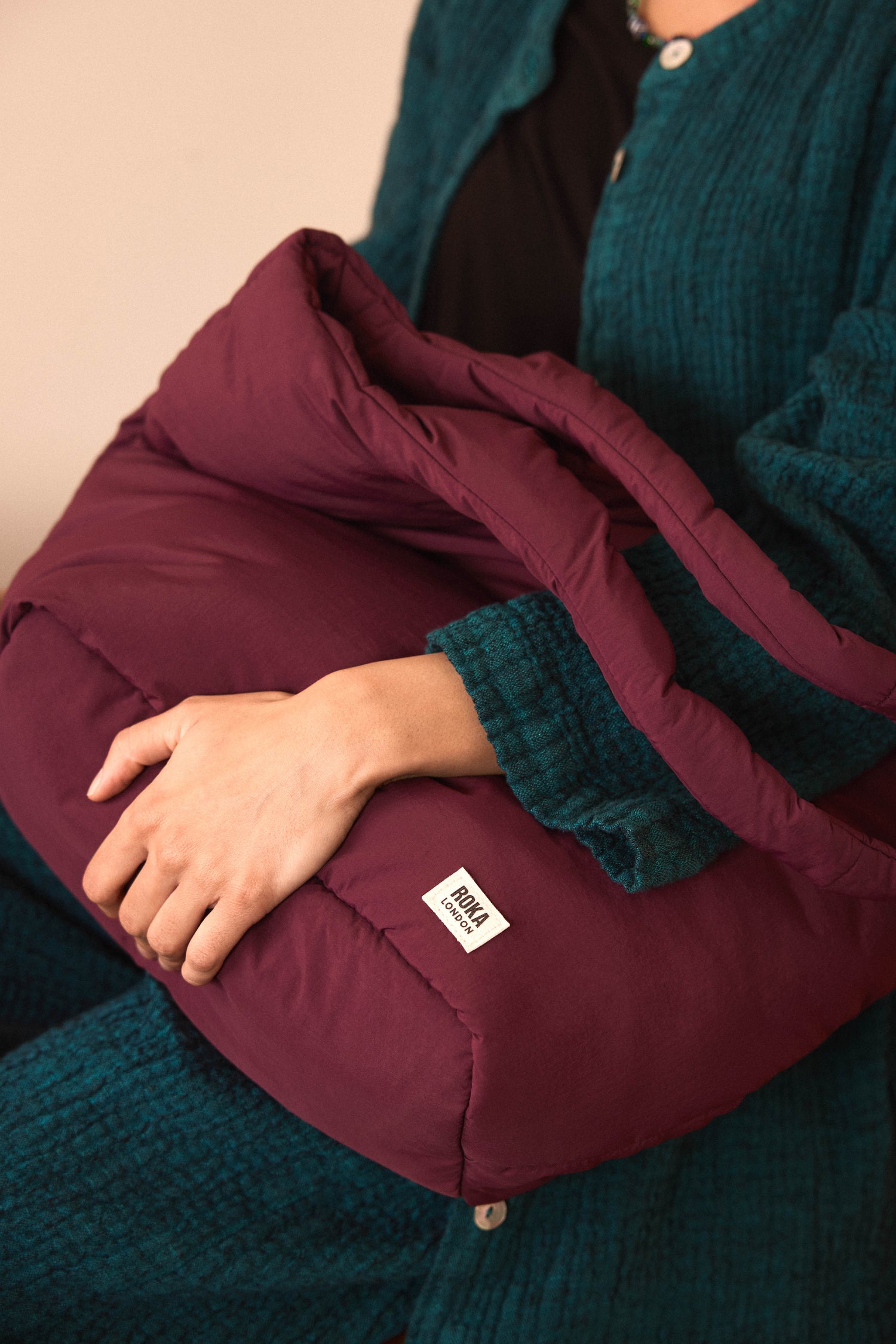 A person in a teal textured outfit holds a maroon quilted pillow or blanket with a visible white “HOMA” label, reminiscent of the cozy padding found in an Oxford Merlot Shoulder Bag, which features a weather-resistant finish and a dedicated laptop sleeve.
