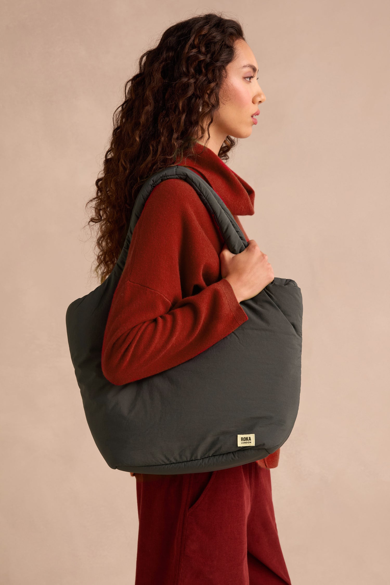 A woman with long curly hair wears a red outfit and carries a large weather-resistant Oxford Raven Shoulder Bag, complete with a padded laptop sleeve.