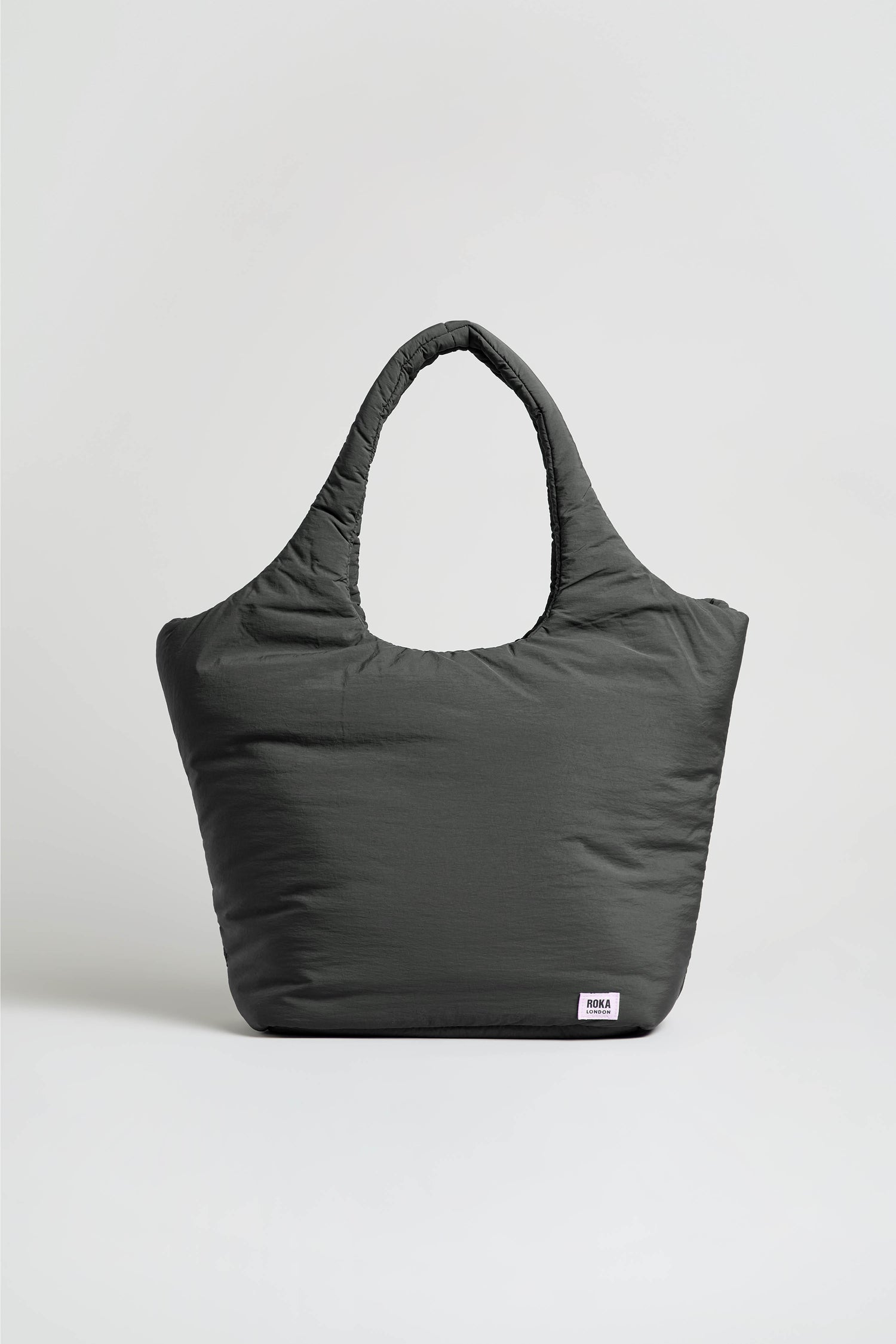 The Oxford Raven Shoulder Bag, shown in a generous size and deep gray shade, features two handles and is set against a simple, light-colored background. Made from weather-resistant material, this tote includes a padded laptop sleeve inside and is finished with a small white rectangular label sewn near the bottom right corner.