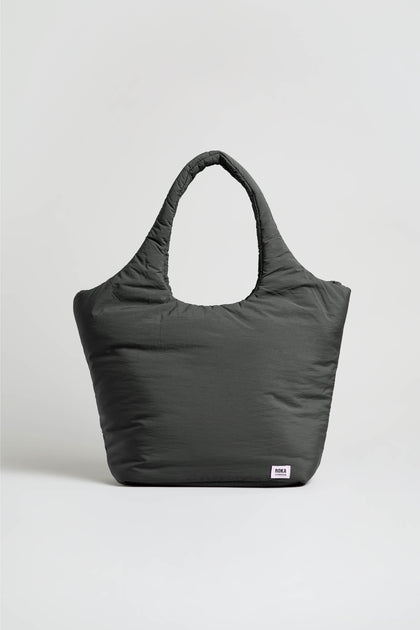 The Oxford Raven Shoulder Bag, shown in a generous size and deep gray shade, features two handles and is set against a simple, light-colored background. Made from weather-resistant material, this tote includes a padded laptop sleeve inside and is finished with a small white rectangular label sewn near the bottom right corner.