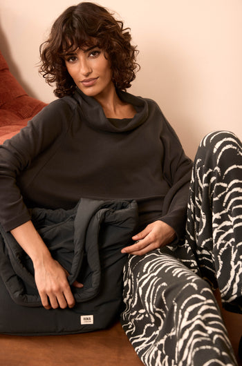 A woman with curly hair sits on a couch, wearing a dark cowl-neck sweater and black-and-white patterned pants, holding the Oxford Raven Shoulder Bag—with weather-resistant fabric and a padded laptop sleeve—in her lap.