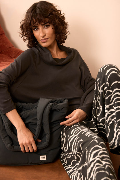 A woman with curly hair sits on a couch, wearing a dark cowl-neck sweater and black-and-white patterned pants, holding the Oxford Raven Shoulder Bag—with weather-resistant fabric and a padded laptop sleeve—in her lap.