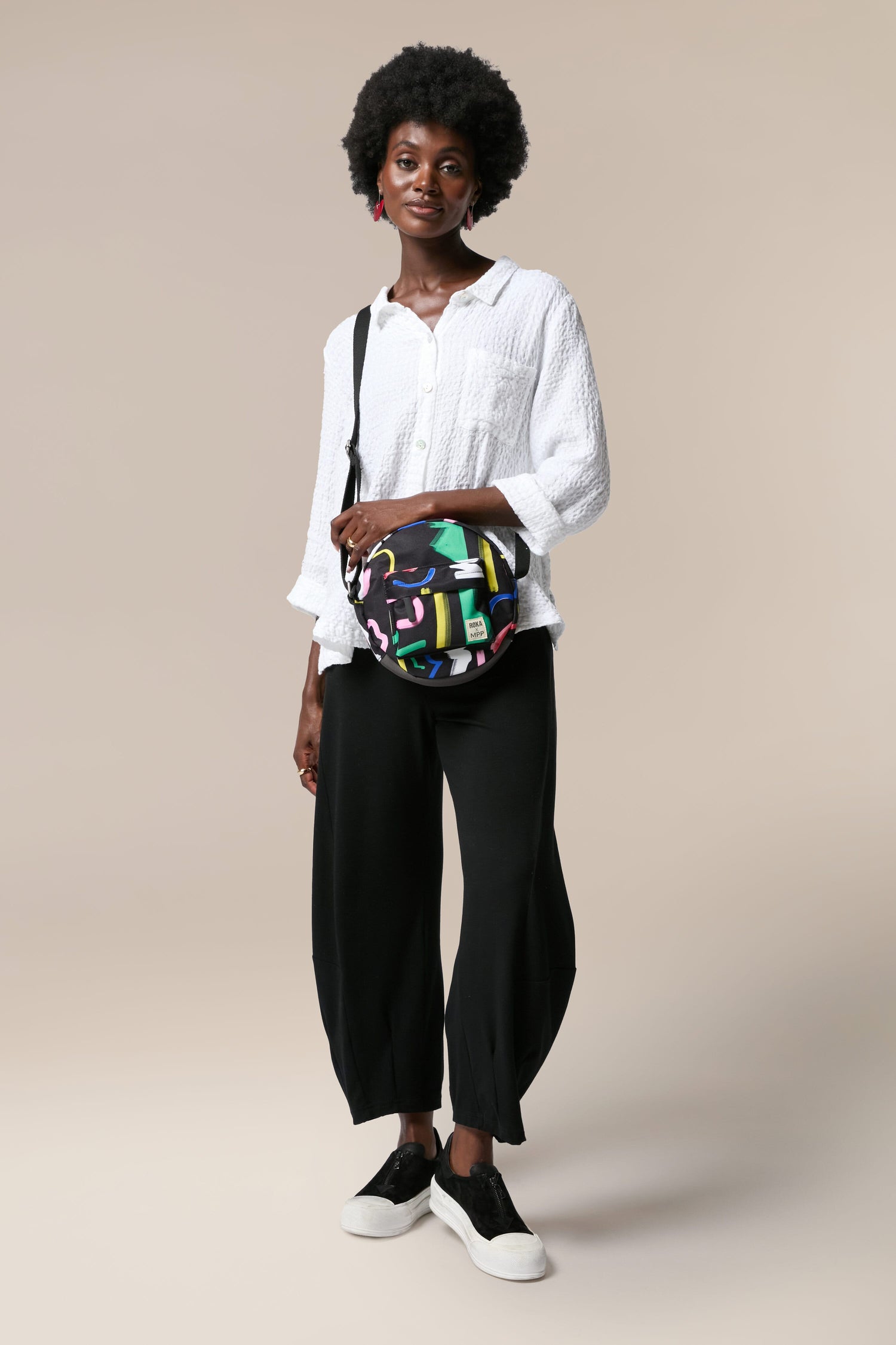 A vegan woman sporting black pants and a weather-resistant, Recycled Scribble Print Paddington Bag in vibrant colors.