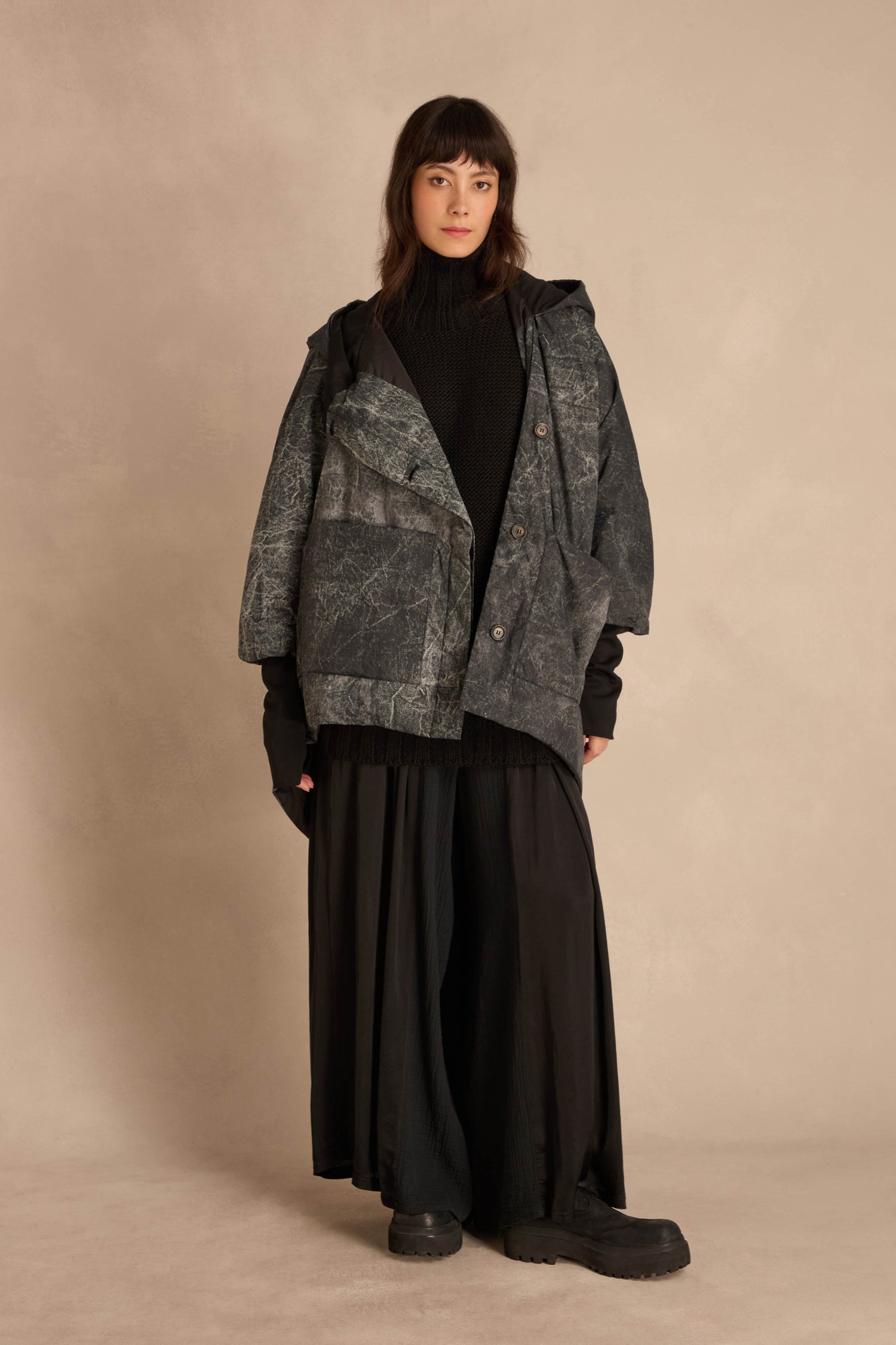 A person wears a dark, textured, asymmetric Asymmetric Hooded Puffer Jacket over a black sweater and long skirt.
