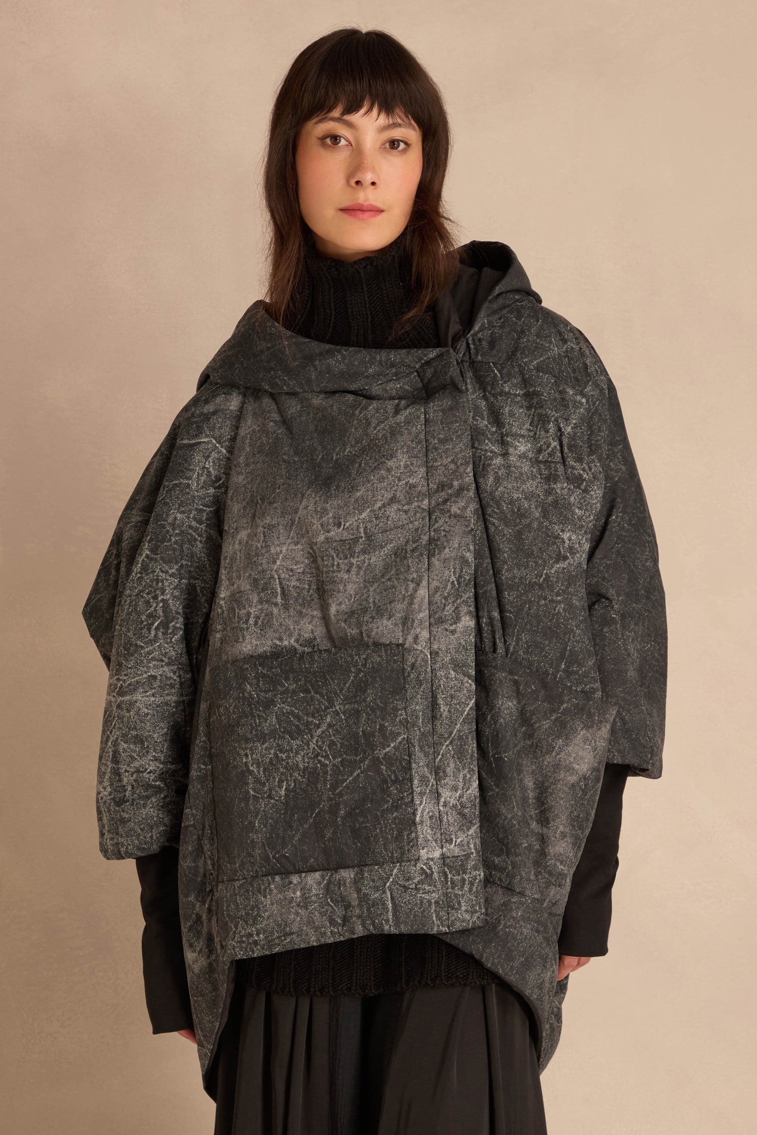 A person wearing an Asymmetric Hooded Puffer Jacket with an oversized, distressed, dark gray patterned finish and asymmetric front closure over a black turtleneck and skirt.