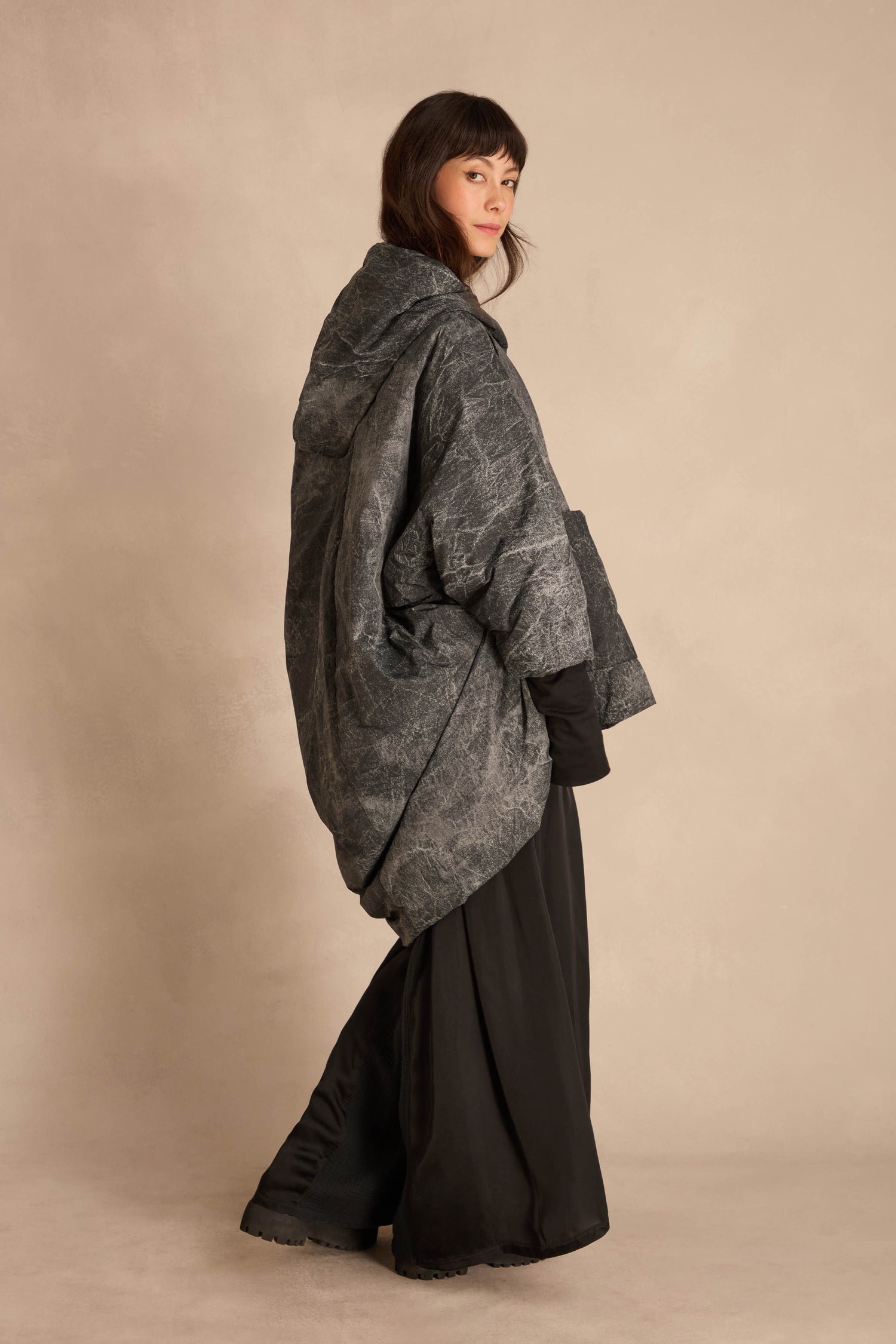 A person wearing an oversized, hooded, grey-patterned Asymmetric Hooded Puffer Jacket over a black skirt.