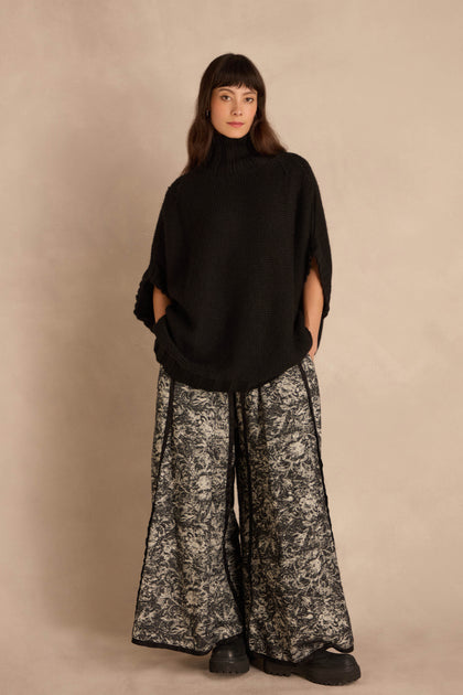 A model wears an oversized, black Black High Neck Knit poncho over wide-leg patterned trousers.