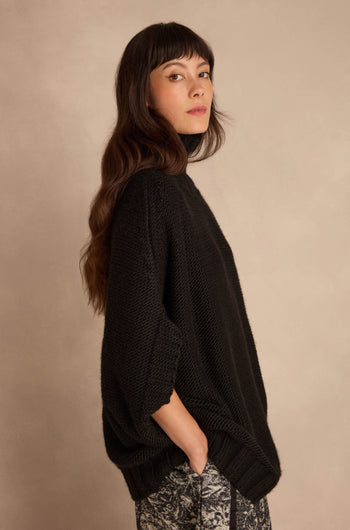A woman wearing a Black High Neck Knit sweater with an oversized, high-neck fit and black three-quarter sleeves.