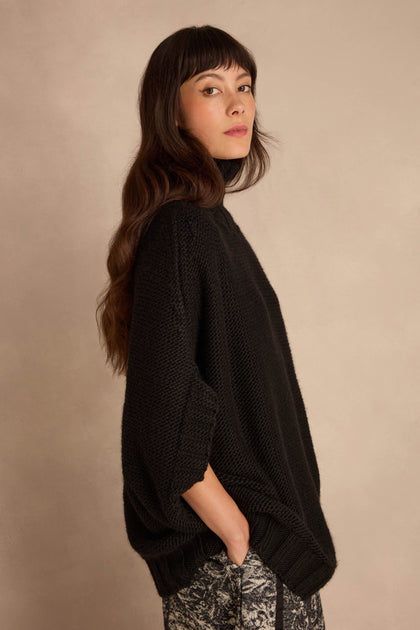 A woman wearing a Black High Neck Knit sweater with an oversized, high-neck fit and black three-quarter sleeves.