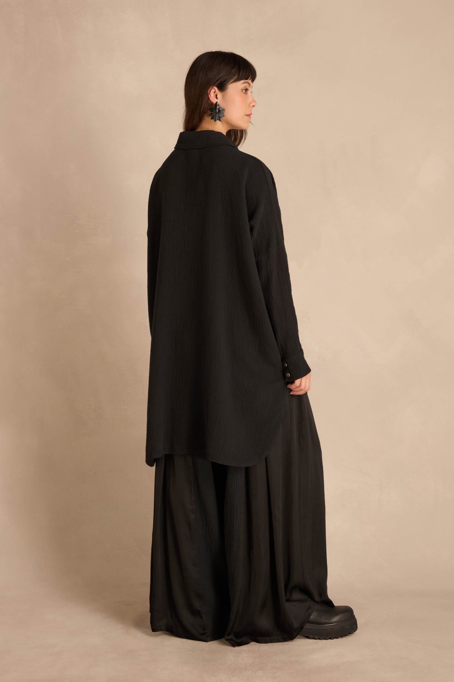 A person seen from behind wears a Black Pocket Satin Shirt in oversized, crinkled cotton with satin patch pockets, paired with long, wide-leg black trousers.