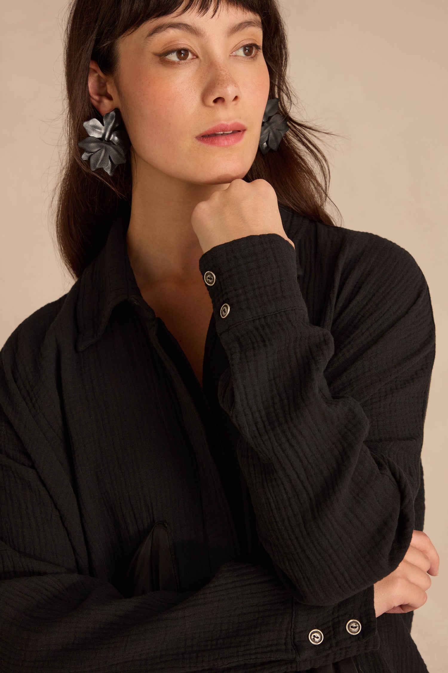 A person wearing an oversized Black Pocket Satin Shirt in black crinkled cotton with textured detailing and large, dark floral-shaped earrings.