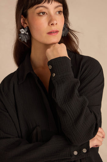 A person wearing an oversized Black Pocket Satin Shirt in black crinkled cotton with textured detailing and large, dark floral-shaped earrings.