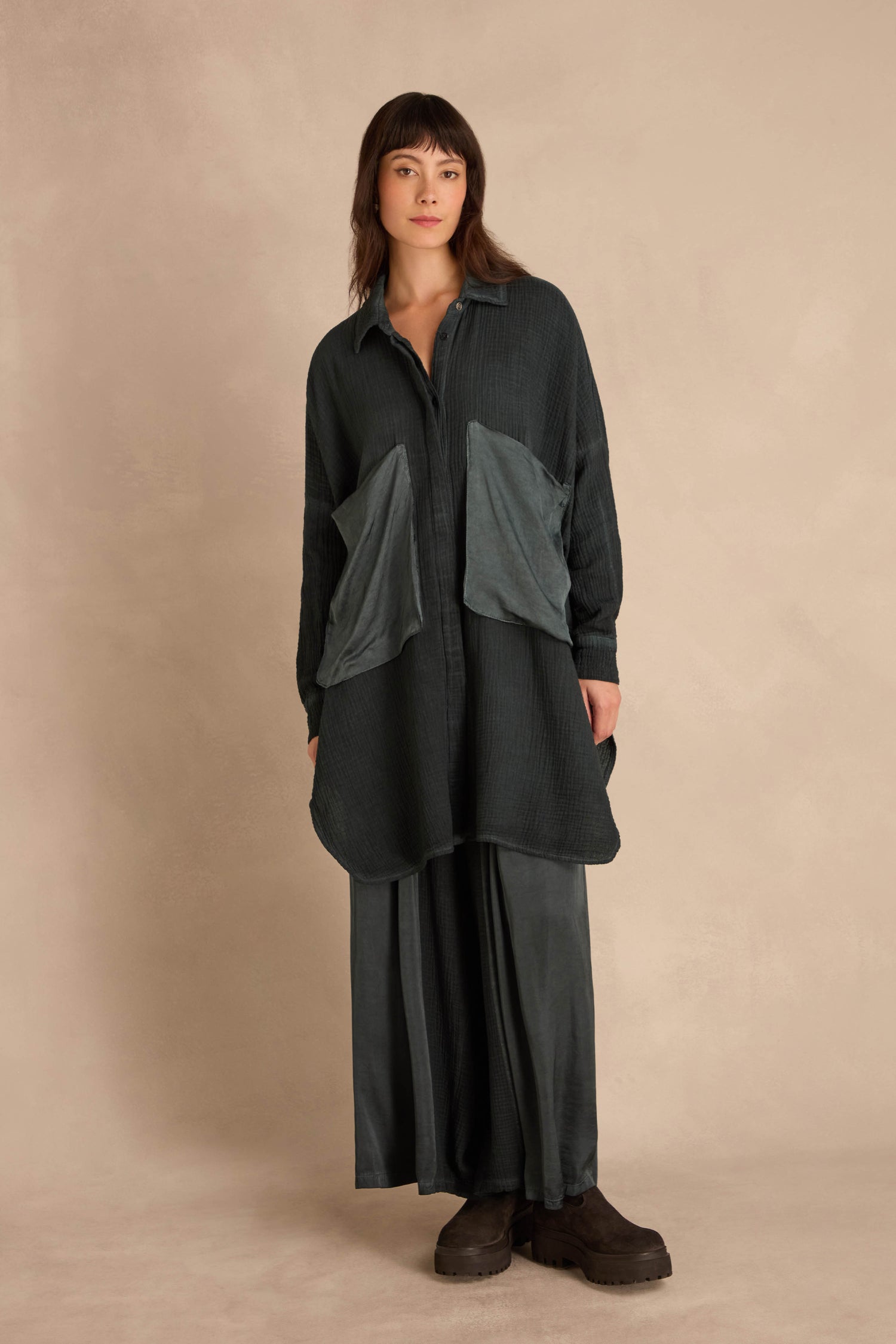 A person wearing an oversized, crinkled Petrol Pocket Satin shirt with large front pockets and a matching long skirt, paired with dark platform boots.
