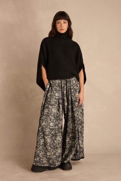 A person wears a black turtleneck poncho and Frayed Hem Wide Leg Trousers in a black-and-white floral pattern with frayed hems.
