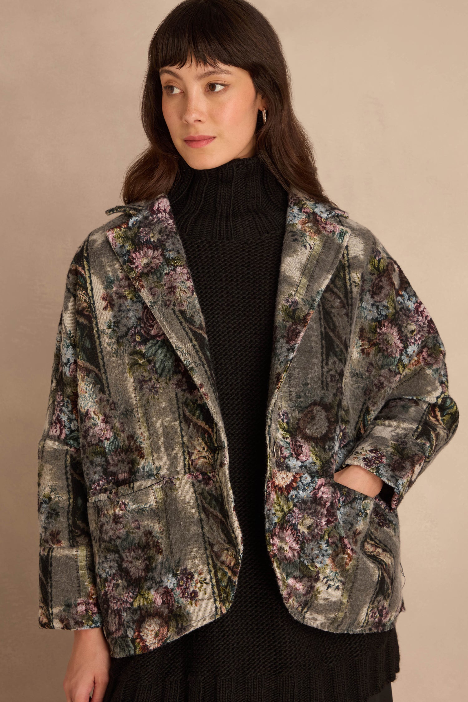 A person wears a charcoal black turtleneck under a boxy, open Tapestry Jacket featuring a muted grey floral tapestry pattern.