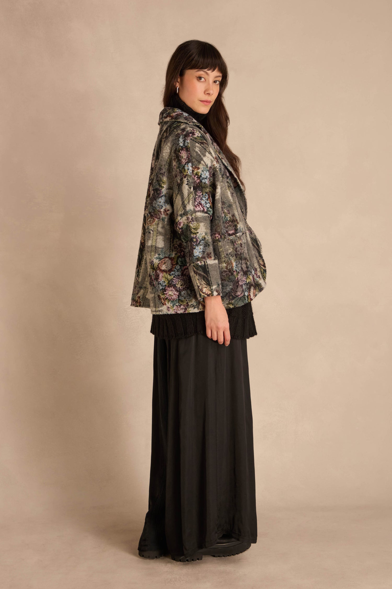 A person wears a cropped Tapestry Jacket featuring a muted floral tapestry pattern over a black turtleneck and a long, pleated black skirt.