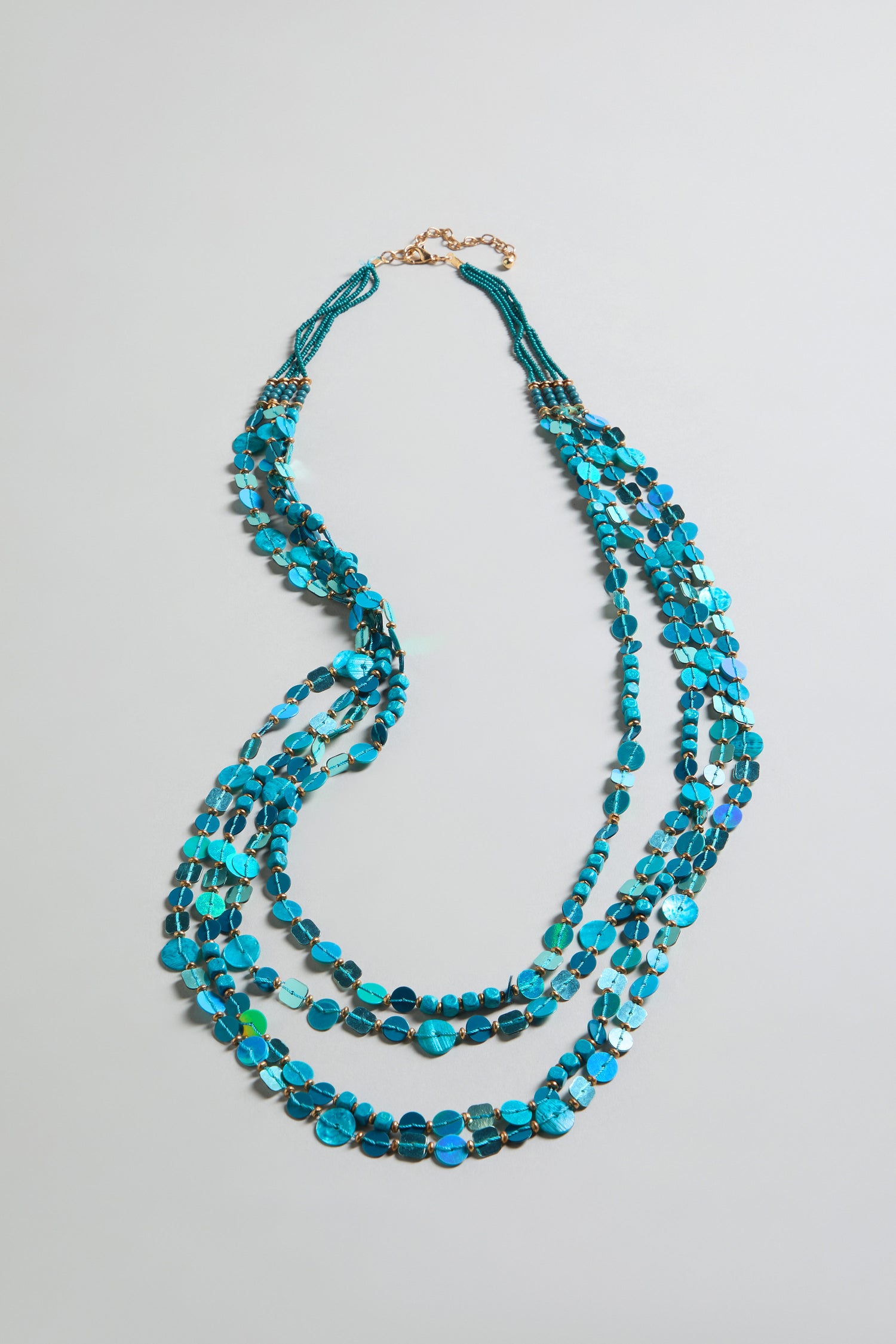 The Four Strand Sequin Necklace features irregularly shaped turquoise beads and a gold adjustable lobster clasp, displayed on a light gray background.