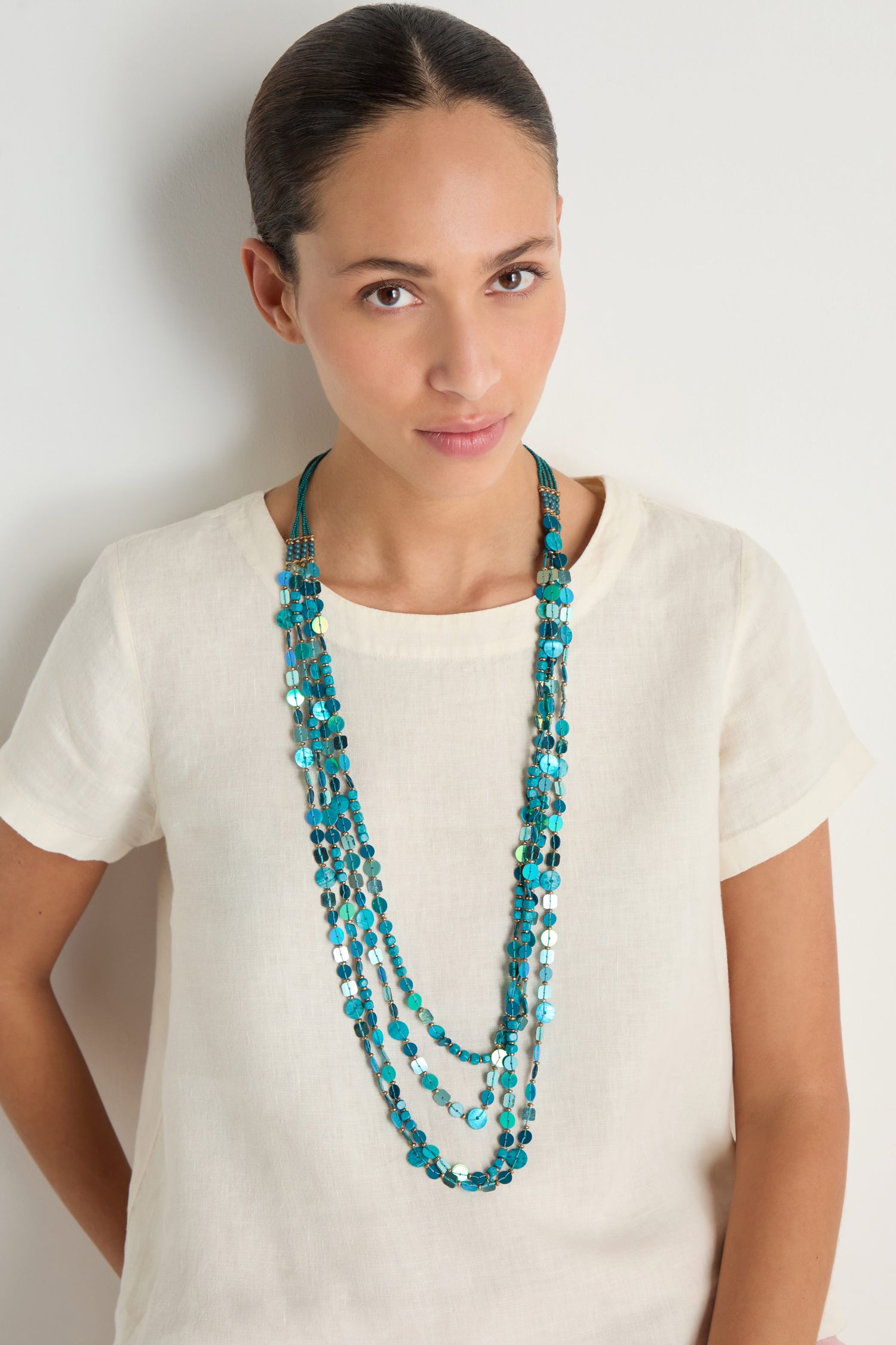 A woman in a cream short-sleeve top and the Four Strand Sequin Necklace with blue beads stands against a plain white background.