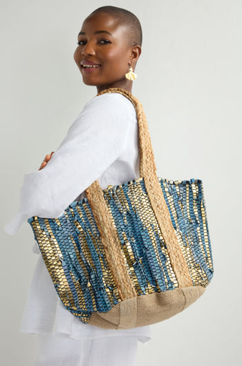 A person in a white outfit smiles, holding a large Blue Upcycled Leather Tote Bag with straw handles, showcasing sustainable craftsmanship.