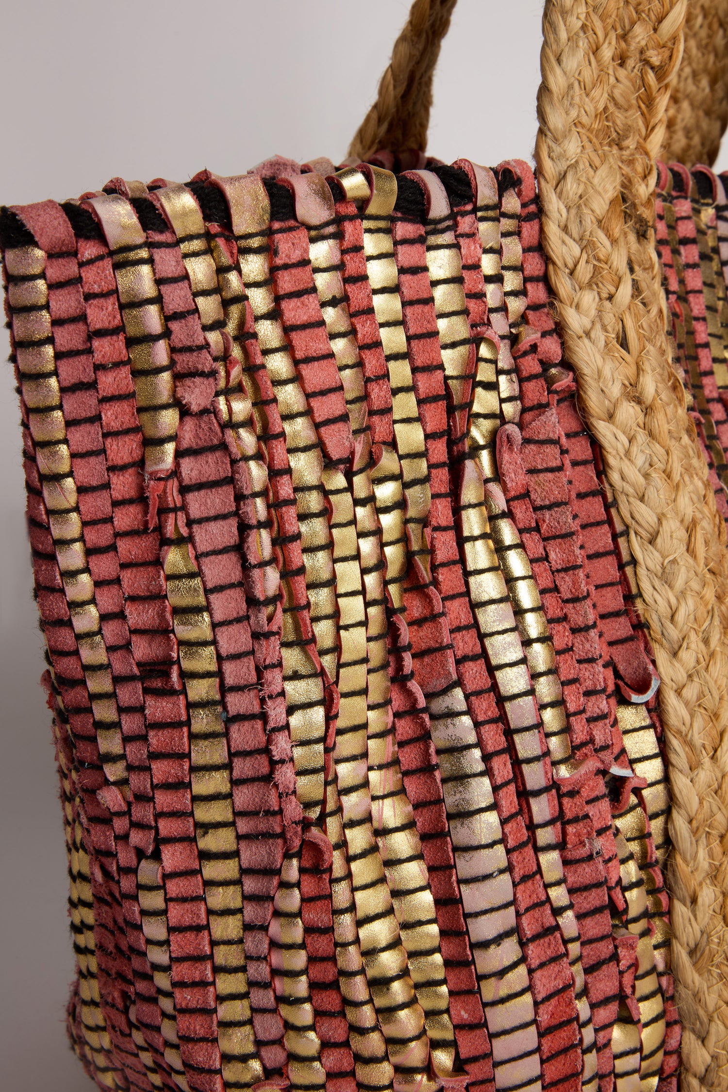 The Rose Upcycled Leather Tote Bag, handmade in India, features woven pink and gold strips with thick braided handles, showcasing textured patterns and sustainable craftsmanship that celebrate artisanal skill.