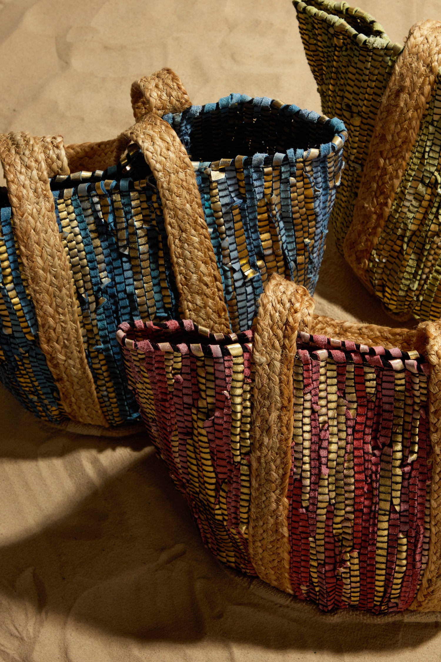 A trio of Rose Upcycled Leather Tote Bags, vibrant in blue, green, and red stripes, sits on sandy terrain. Each tote boasts robust textured handles and highlights eco-friendly artistry.