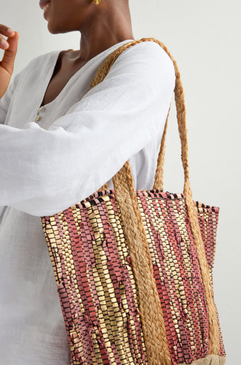 A person in a white long-sleeve shirt holds the Rose Upcycled Leather Tote Bag with earth-toned patterns and large rope handles. Handmade in India, its intricate design showcases sustainable craftsmanship as they face away from the camera.