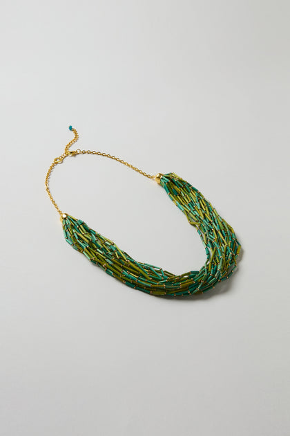 The Multi Strand Grass Bead Necklace features green grass beads, gold accents, a gold-look clasp chain, and an adjustable closure. Shown on a light gray background.