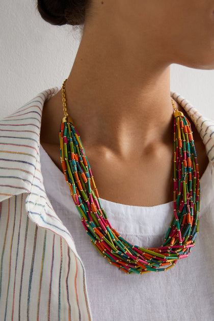 A person wears the Multi Strand Grass Bead Necklace, featuring a polished gold-look clasp chain, styled with a white top and a white shirt with thin colorful stripes.