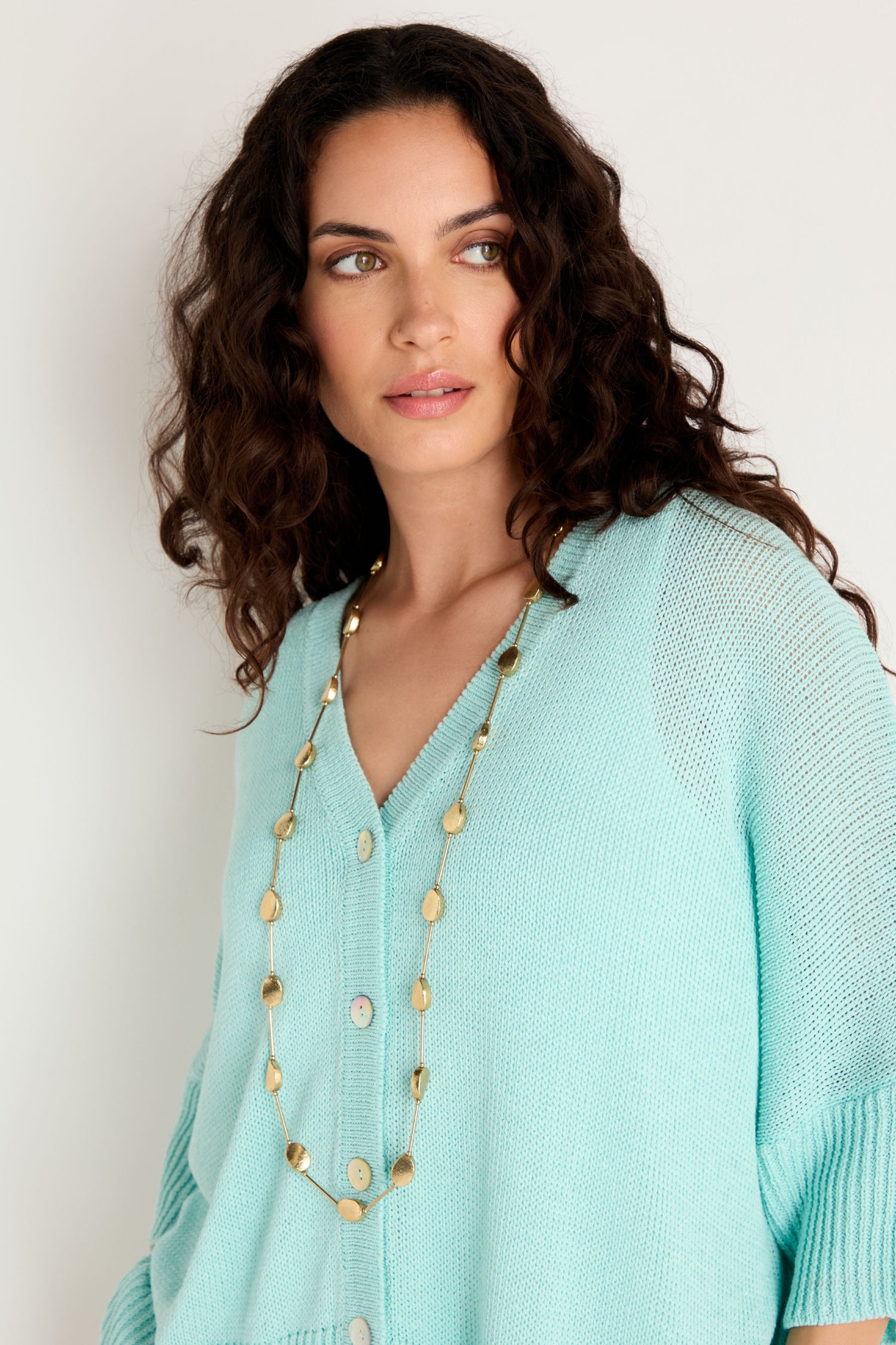 A woman with curly dark hair wears a light blue knit cardigan and the Long Oval Disc Necklace, featuring textured oval discs, as she looks slightly to the side against a plain background.