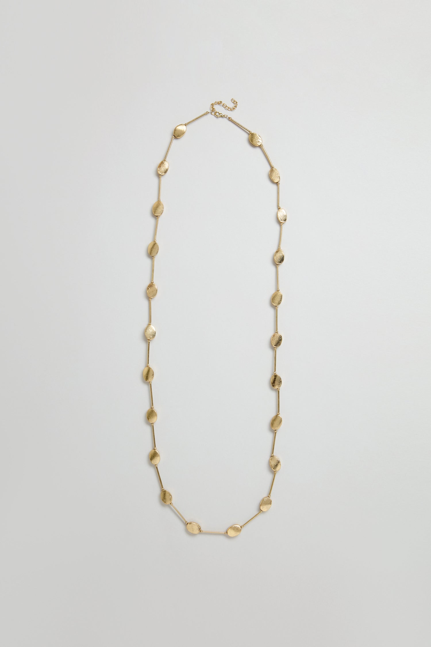 The Long Oval Disc Necklace features textured oval discs evenly spaced along its metallic chain, showcased on a plain light background.